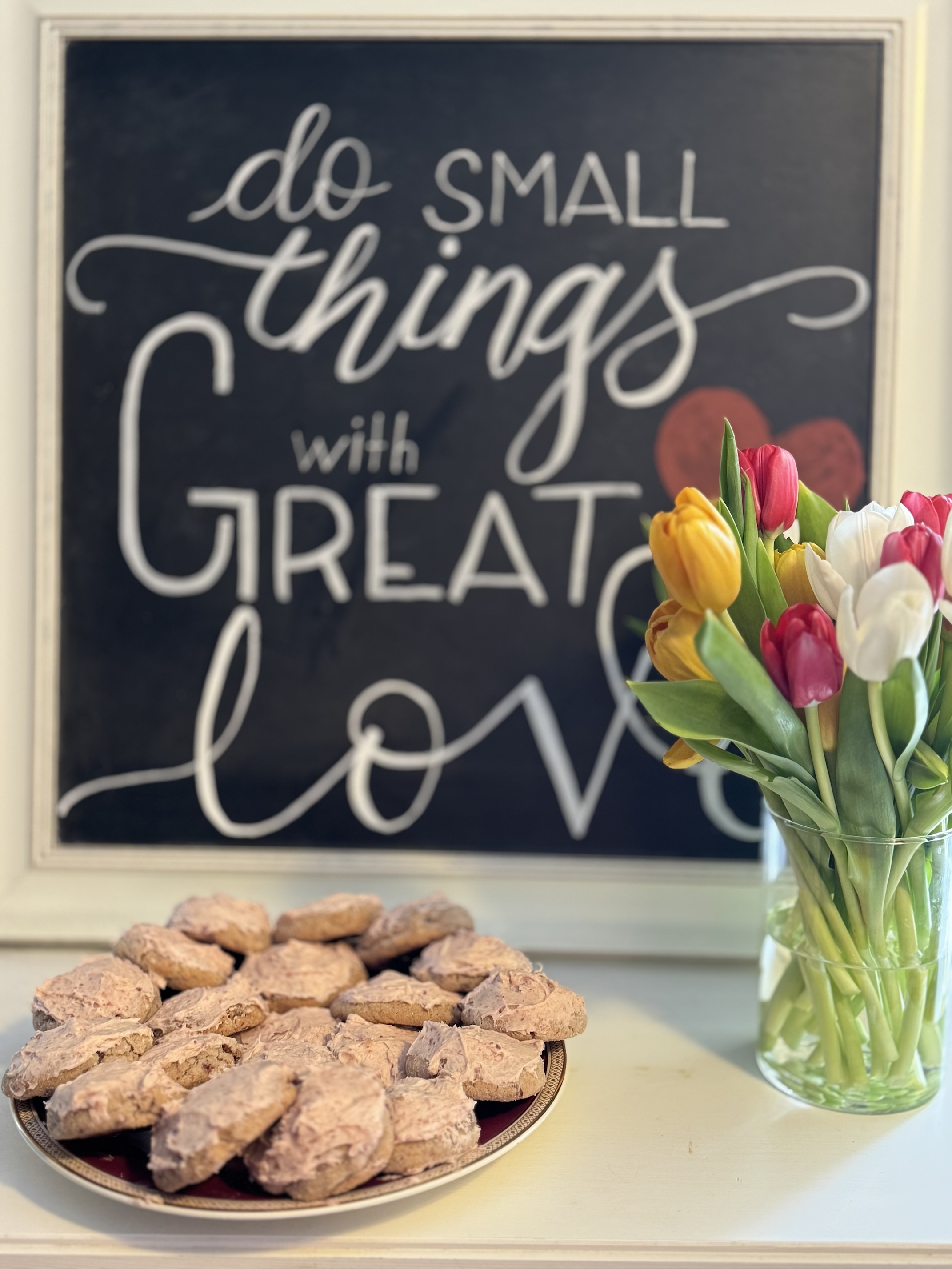 Plate of cookies in the left foreground. Cookies are covered in a thick layer of pink strawberry icing. Right foreground is a vase of a dozen red and yellow tulips. Background is a chalkboard with calligraphy that says 'Do small things with great love.'